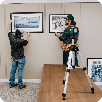 Two Gardner employees hanging framed pictures on a wall in an office using precise laser levels