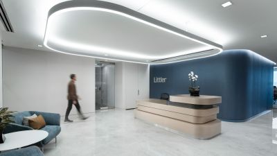 Modern office reception area with a curved wood reception desk, sculptural overhead lighting, blue accent wall, lounge seating, and an employee walking through the lobby.