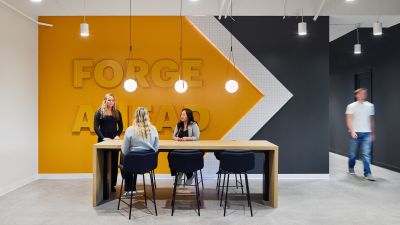 Employees collaborate around a high-top meeting table in a modern office featuring a bold yellow accent wall with "Forge Ahead" signage, pendant lighting, and contemporary finishes.