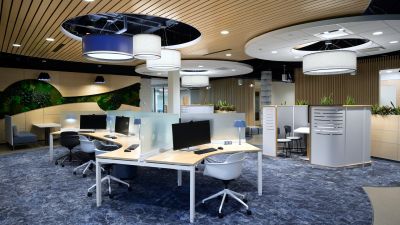 Modern open office workspace with shared desks, semi-private work pods, biophilic design elements, circular ceiling features, and collaborative seating areas.