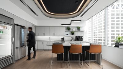 Modern office caf&eacute; with a large island, white cabinetry, floor-to-ceiling windows, contemporary lighting, and an employee using the beverage station.