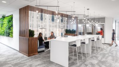 Modern commercial office collaboration space with white communal tables, built-in meeting booths, wood accents, a living green wall, and employees meeting and working throughout the space.