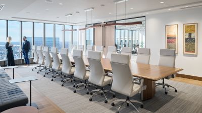 Large executive boardroom with a long conference table, white leather chairs, floor-to-ceiling windows, city views, and two employees talking near the windows.