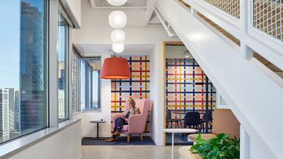 Modern office lounge beneath a staircase with floor-to-ceiling windows, colorful patterned walls, a private nook with an employee seated in a lounge chair using a mobile device.