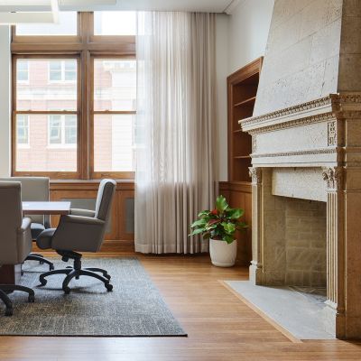 Historic meeting room with ornate fireplace on the right, wooden floors and windows, and a center table.