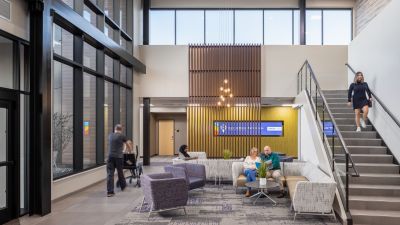 Healthcare facility lobby with comfortable waiting areas, a grand staircase, floor-to-ceiling windows, and patients and visitors gathering in a bright, welcoming environment.
