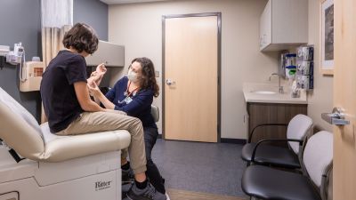 Healthcare exam room where a clinician wearing a face mask examines a patient's arm, featuring an exam table, sink, storage cabinetry, and a clean, patient-centered clinical environment.