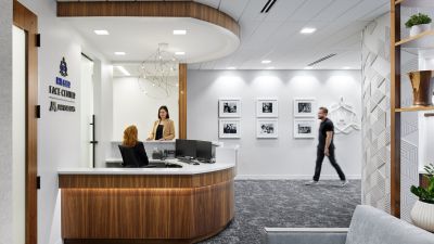 Modern healthcare clinic reception area with a curved wood reception desk, contemporary lighting, welcoming seating, and staff assisting patients in a bright, hospitality-inspired environment.