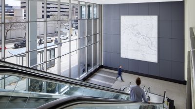 Bright building lobby with floor-to-ceiling windows, escalators, and a large illuminated stone feature wall.