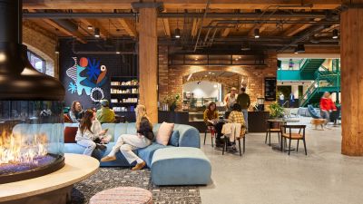 People gather in a renovated historic atrium featuring exposed timber beams, brick arches, a coffee shop, abstract mural, and lounge seating.