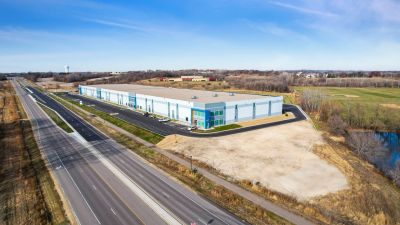 Aerial view of a newly completed industrial warehouse with office entrances, parking lots, and truck access, situated along a major roadway with surrounding commercial development and open green space.