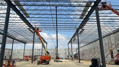 Structural steel framing rises inside a tilt-up industrial warehouse under construction as workers on boom lifts install roof decking above concrete wall panels supported by temporary braces.