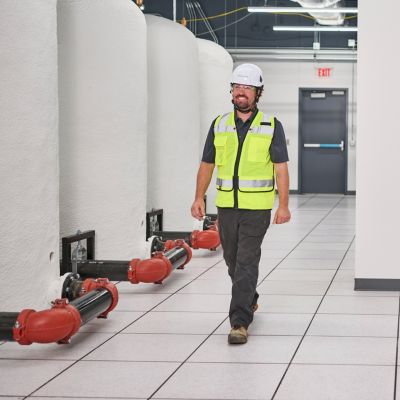 Construction professional wearing a hard hat, safety glasses, and high-visibility vest walking through a mission-critical facility with insulated tanks and piping.