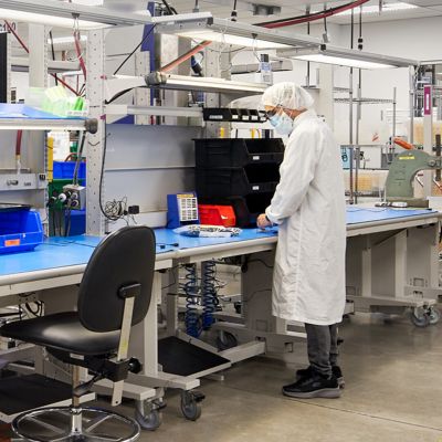 Technician wearing cleanroom attire and a face mask assembling components at a manufacturing workstation in a science and technology facility equipped with organized workstations and specialized production equipment.