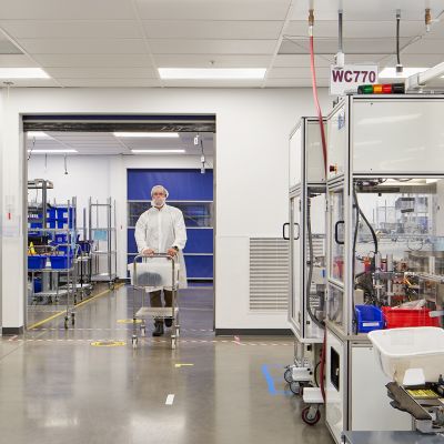 Clean room manufacturing space with automated production equipment and a technician wearing full cleanroom attire while transporting materials, highlighting a controlled environment for precision manufacturing.