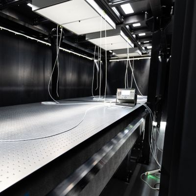 Precision optics laboratory featuring a large vibration-isolated testing table, overhead lighting panels, suspended cables, and specialized measurement equipment for research and advanced technology development.