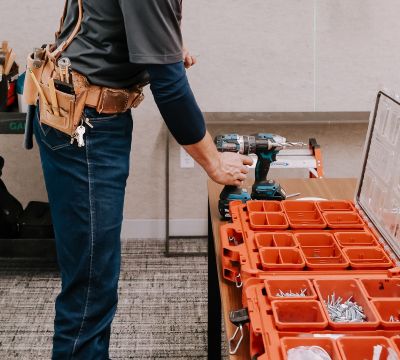 Technician wearing a tool belt organizes drills and hardware from a portable parts organizer while preparing to complete post-construction service work in a finished commercial office space.