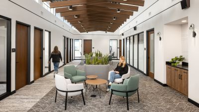 Office corridor with wood ceiling beams, a seating area with green and white chairs, and a woman seated using her phone.