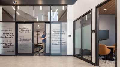 Hallway with frosted glass doors printed with cooperative principles, a meeting room visible through the glass, and a man walking through.