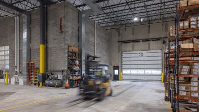 Interior of a vehicle service bay with concrete block walls, a large overhead door numbered "20," storage shelving, and a forklift in motion.