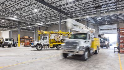 Interior of a truck garage with utility trucks, including a bucket truck with a Terex crane, parked near storage shelves and an open bay door.