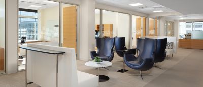 A modern office lounge with blue high-back chairs, white benches, and glass-walled private offices in the background.