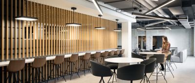 A sleek lounge with wooden slat walls, bar stools, and geometric ceiling lights where a woman sits at a table.