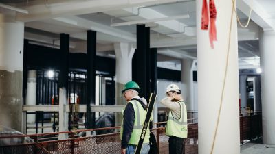 Two construction workers in hard hats and safety vests survey an under-construction interior with exposed columns and beams.