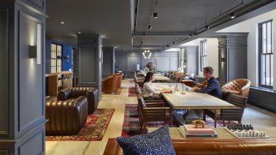 Wood-paneled coworking lounge with tufted leather chairs, patterned rugs, and people working at a long communal table.
