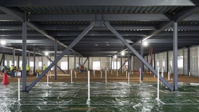 Interior view of exposed steel framing above a dirt subfloor, with workers reviewing plans nearby.