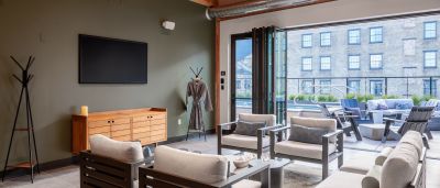 Lounge with grey outdoor-style furniture facing a folding glass door open to a pool deck with adjacent brick building views.