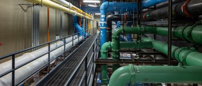 An industrial mechanical room with color-coded pipes (yellow, blue, green, white) running along a catwalk.