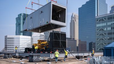 A crane lowers a large gray enclosure onto a rooftop generator installation, with the Minneapolis skyline in the background.