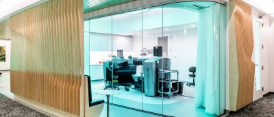 A glass-walled procedure room lit in teal, showing laser equipment and a curtained bay, framed by a wood-slat wall.