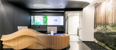 A sculptural wood reception desk faces a video screen, with a forest-themed mural on the adjacent wall.