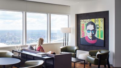 A woman reads on a sofa beside floor-to-ceiling windows, near a colorful pop-art portrait on the wall.