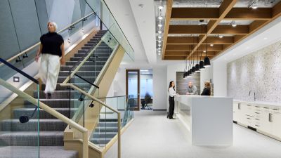 A woman descends a wood and glass staircase near a reception desk where two colleagues greet a visitor.