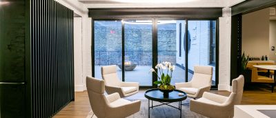 A stylish lounge with beige chairs around a black table and a view of an outdoor fire pit through glass doors.