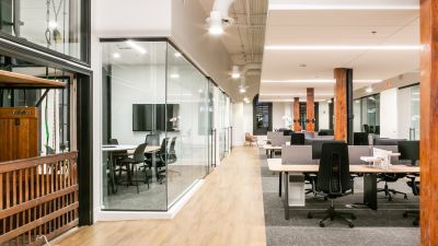 A sleek workspace with glass partitions, modern desks, and exposed ceiling ducts in a bright, open layout.