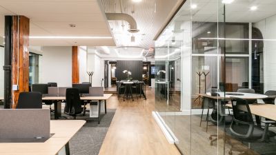 An open office with rows of desks, glass-walled meeting rooms, and wooden columns under bright lighting.